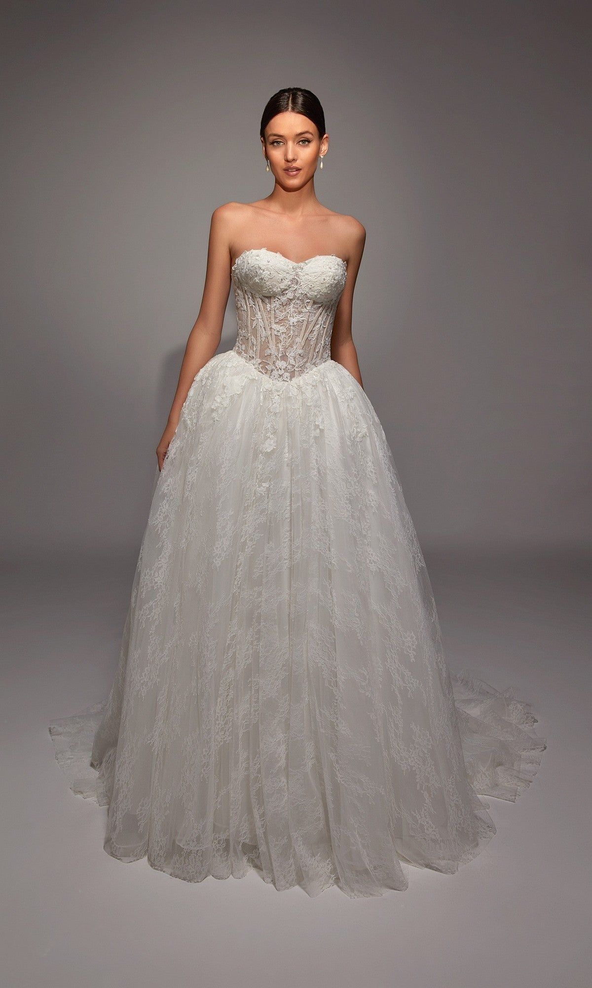 A woman poses in the Long White Dress: Alyce 7163, featuring a strapless, sheer corset bodice and full lace skirt, set against a plain studio backdrop.